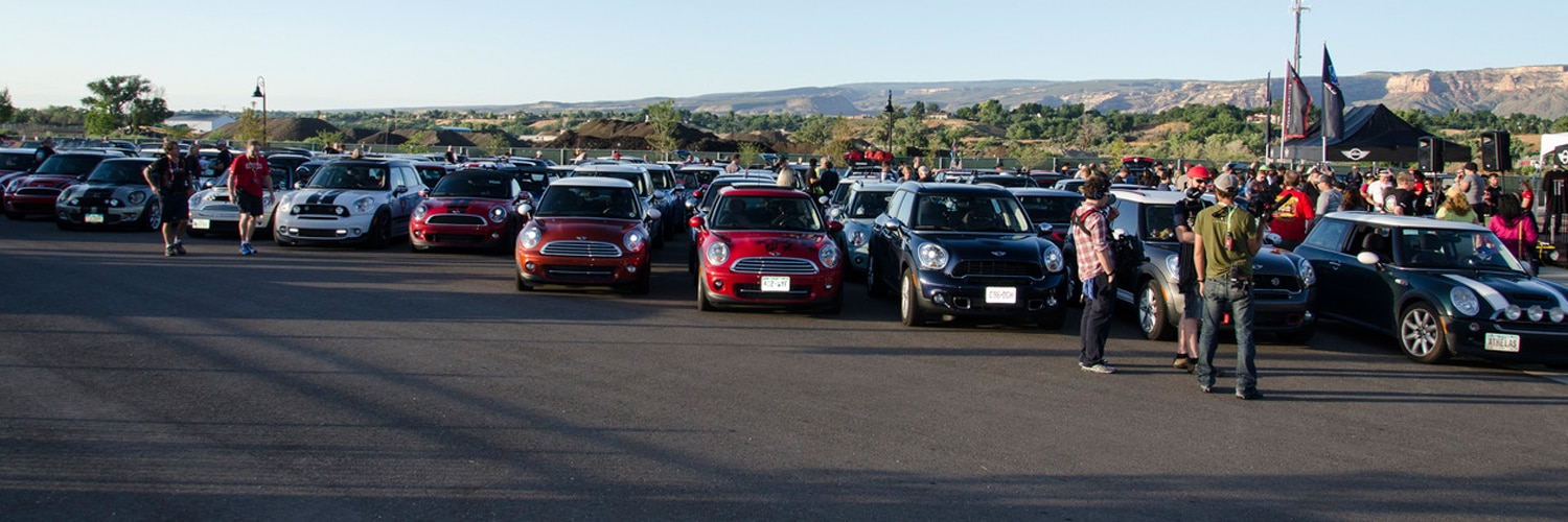 Landscape view of several MINI vehicles of different colors parked in a parking lot, with people walking nearby or standing around in both the foreground and background, on a sunny day with clear skies, trees, mountains, and a MINI tent in the background.